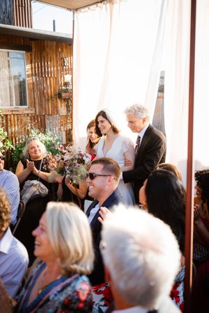 Bride walks down the aisle with escort at outdoor wedding ceremony, guests applauding, rustic wood backdrop.