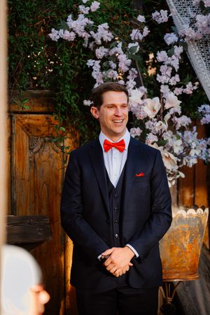 Smiling groom in navy suit with red bow tie and pocket square stands before floral backdrop at wedding ceremony.