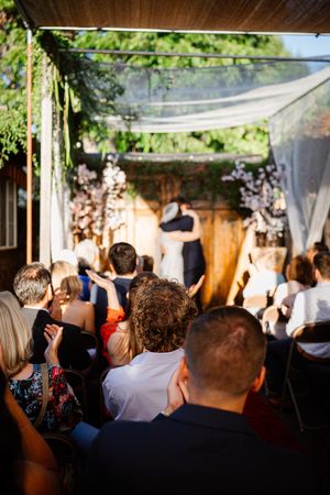 Guests seated at an outdoor wedding ceremony watching couple exchange vows under a floral chuppah.