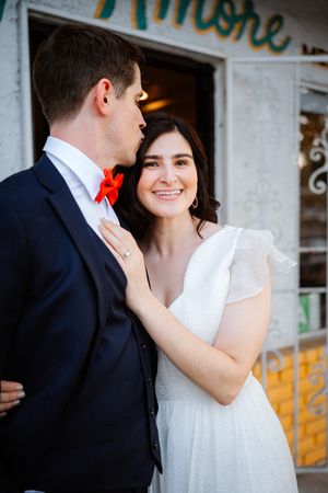 Smiling bride in white dress embraces groom in navy suit with red bow tie outside Amore restaurant.