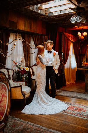 A bride and groom share a romantic moment in an elegant, warmly lit rustic venue with ornate decor and flowing curtains.