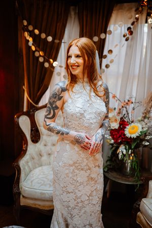 Smiling tattooed bride in lace wedding gown stands before elegant draped backdrop with string lights and floral arrangements.