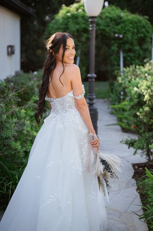 Bride in off-shoulder white lace ballgown with floral appliqués holds a boho bouquet, smiling over her shoulder in a garden.