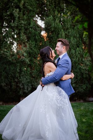 Groom in blue suit lifts bride in lace ballgown wedding dress, laughing together outdoors among tall green trees.