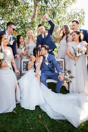 A joyful wedding party surrounds a kissing bride and groom seated on a bench outdoors, bridesmaids in gray gowns holding bouquets.