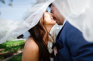 Bride and groom sharing a romantic kiss under a flowing white wedding veil outdoors on their wedding day.