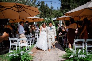 Bride and groom walk down outdoor wedding aisle lined with white chairs and guests under tan patio umbrellas.