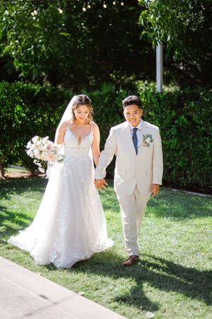 Bride in lace gown and groom in tan suit hold hands walking on green lawn at outdoor wedding.