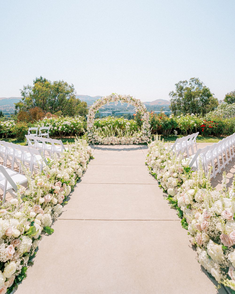 Elegant outdoor wedding ceremony setup with white chairs and floral arrangements along a beachfront stone pathway.