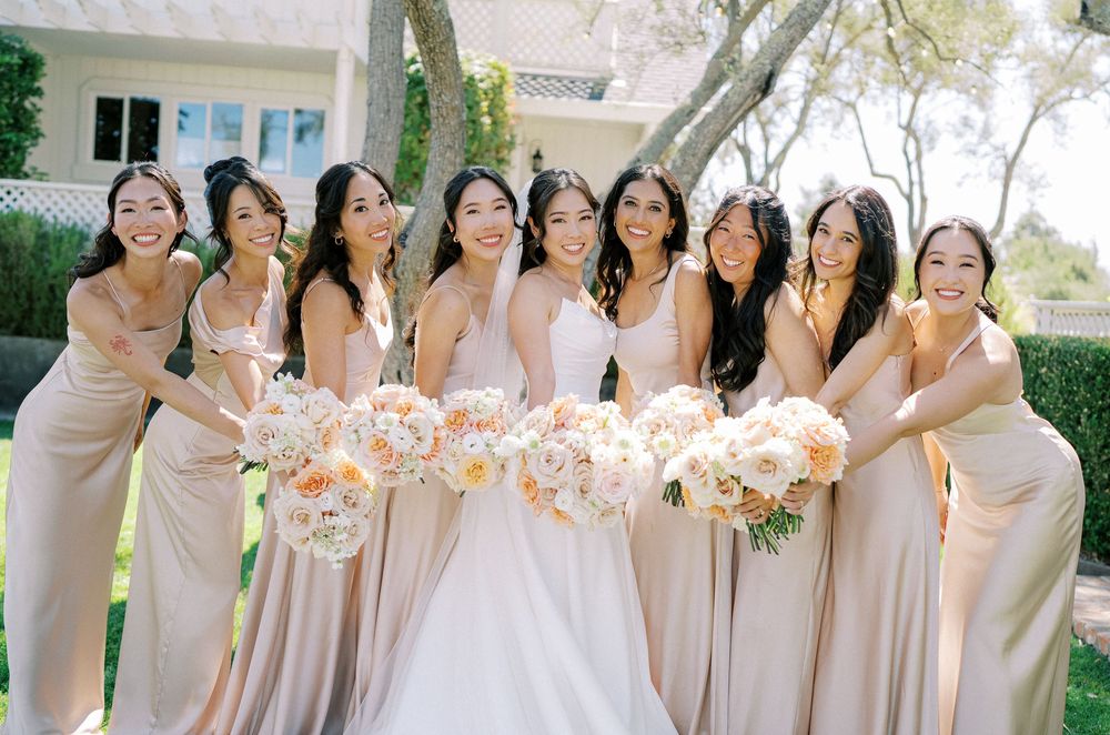A group in blush pink bridesmaid dresses hold matching bouquets while posing together outdoors.
