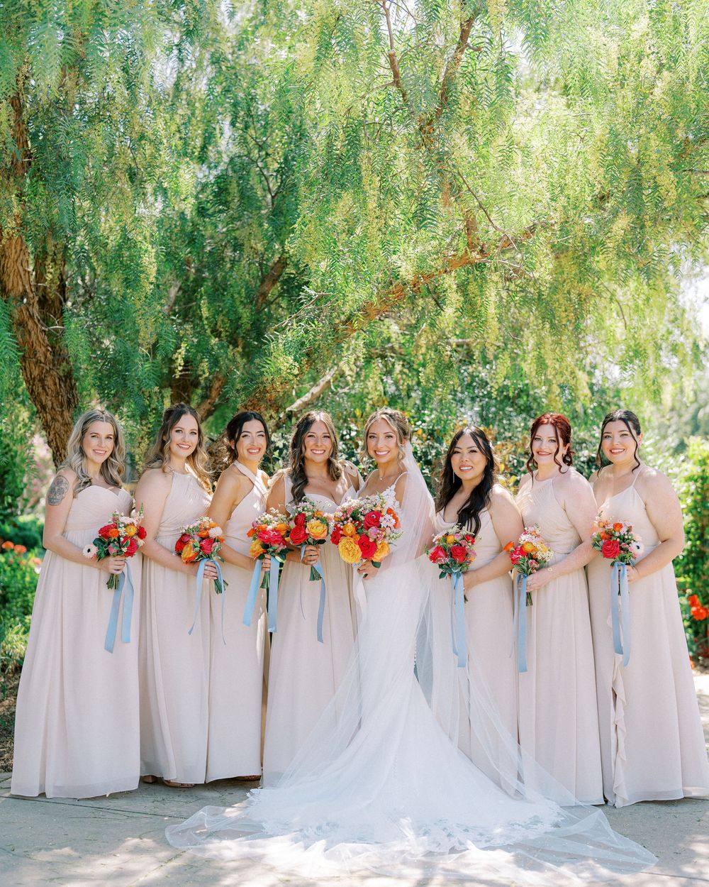 A wedding party in blush pink dresses poses with vibrant orange and red bouquets under weeping willow trees.