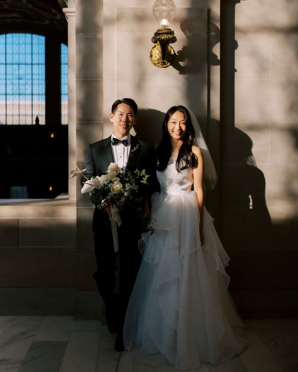 A bride and groom share intimate moments in a dramatic industrial venue with large windows casting natural light.
