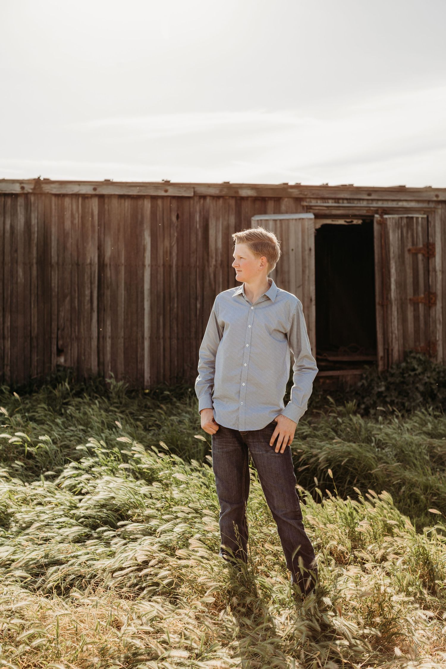 Person standing beside rustic wooden barn in sunlit grassy field.