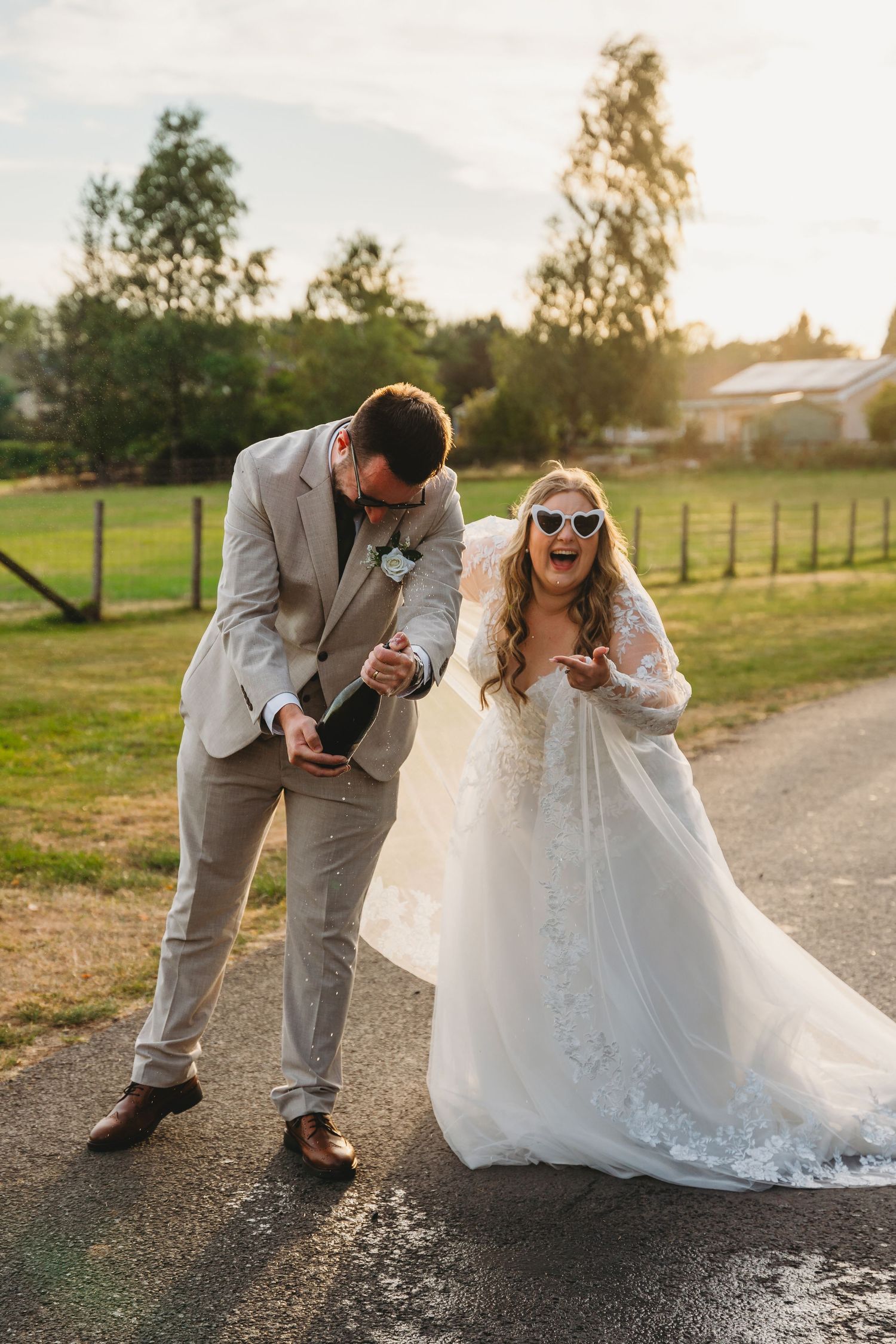 Bride in lace gown and heart sunglasses laughs as groom pops champagne bottle outdoors at golden hour at Canada Lodge South Wales summer