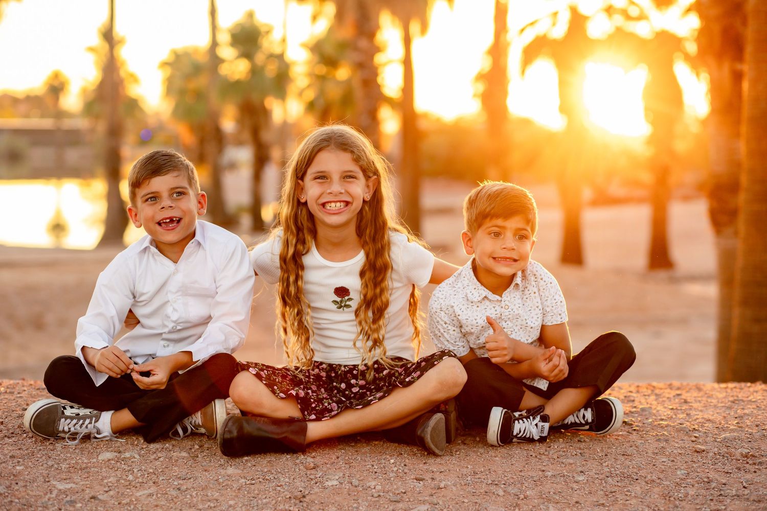 Three happy children sit together outdoors at golden sunset near palm trees.