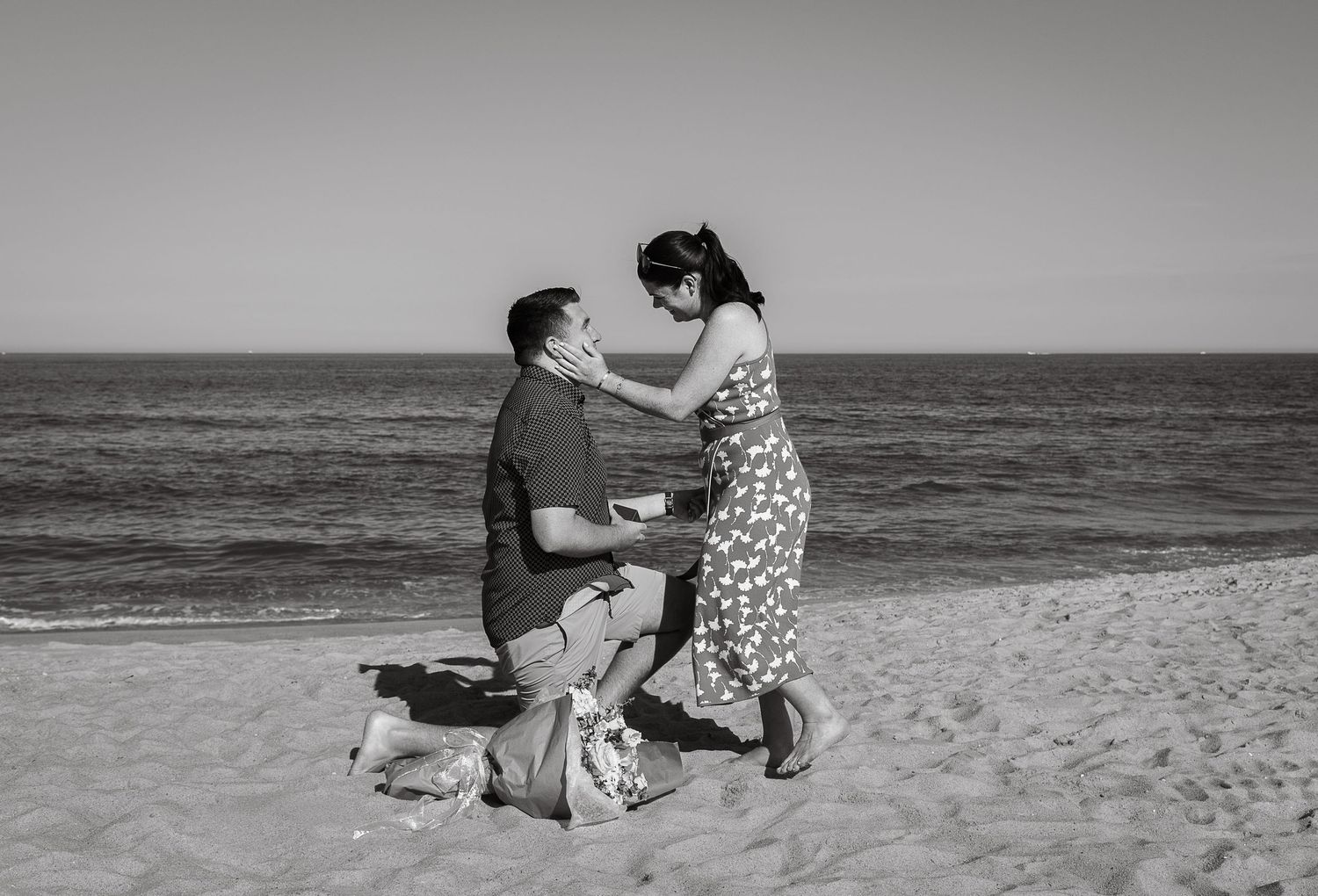 A romantic beach proposal scene showing a couple and dog sitting on sandy shore at sunset.