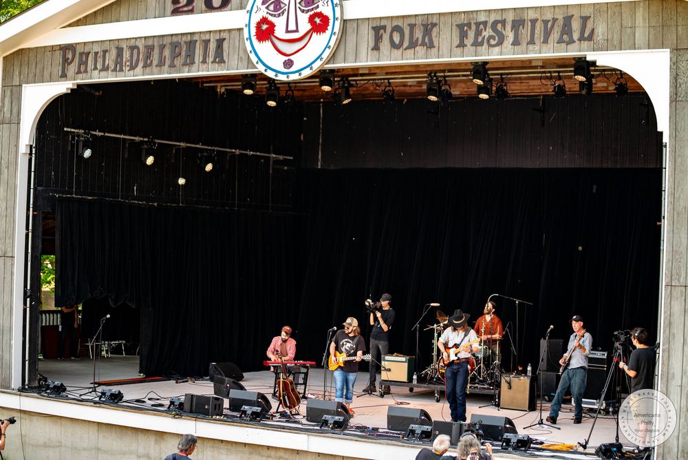 Band performing on stage at the Philadelphia Folk Festival outdoor amphitheater with colorful signage and clown logo.
