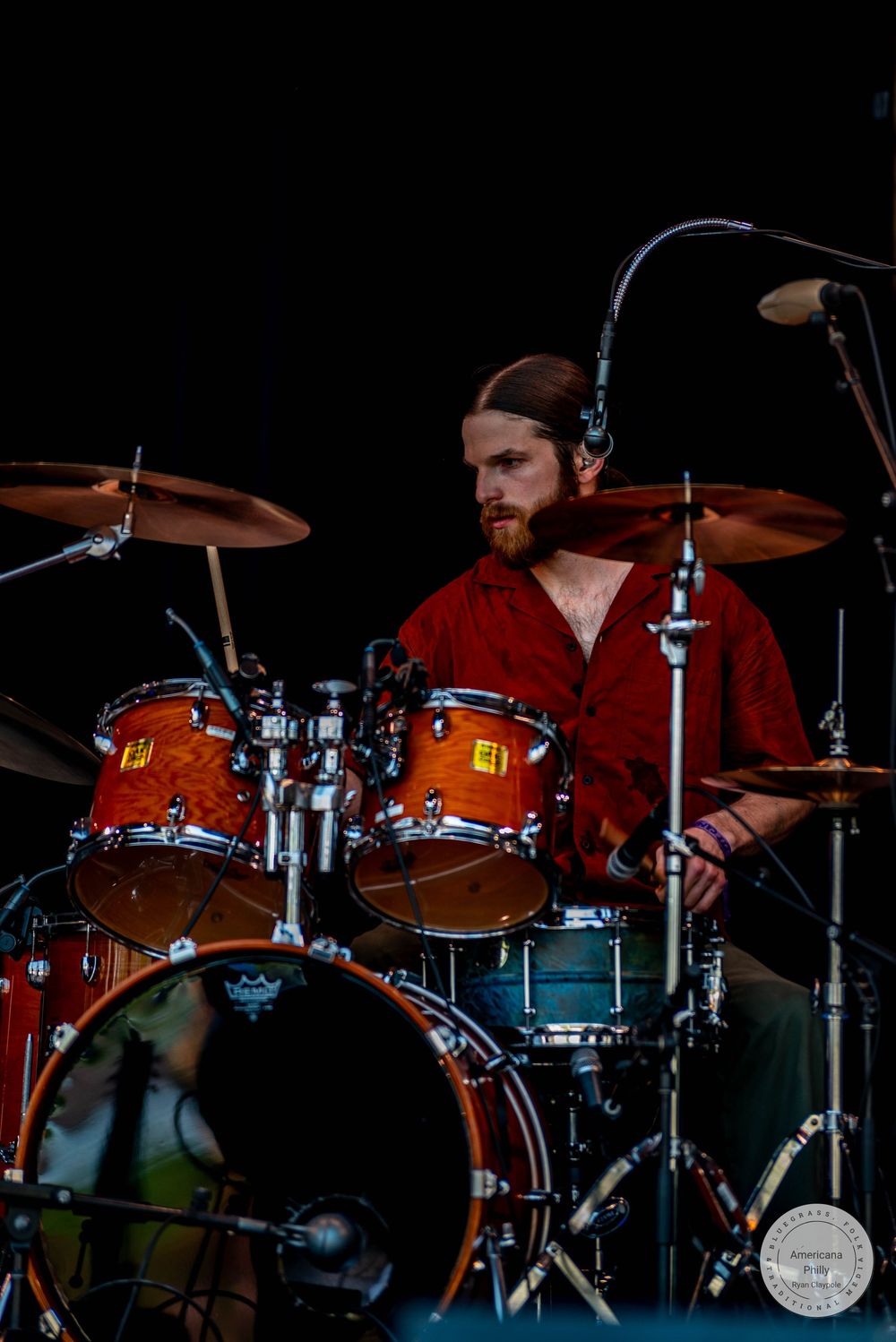 A bearded male drummer in a red shirt performs on stage with a warm-toned drum kit under dramatic lighting.