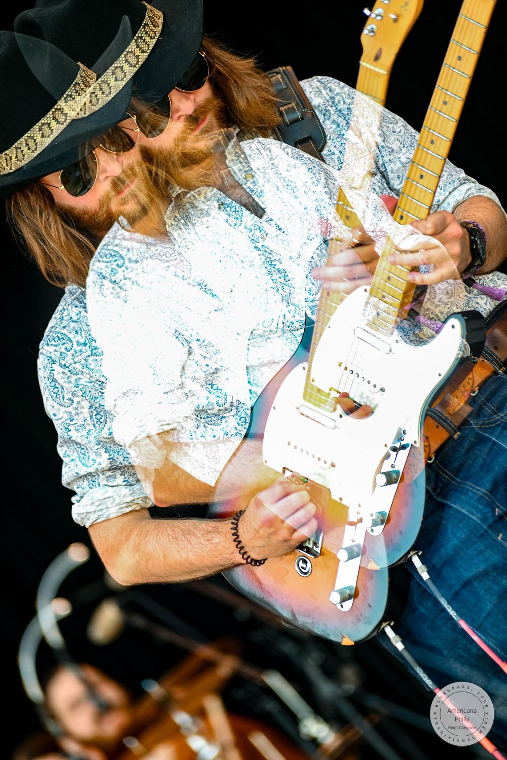 Guitarist in black hat plays a worn Fender Telecaster on stage, wearing a floral shirt with a bracelet.