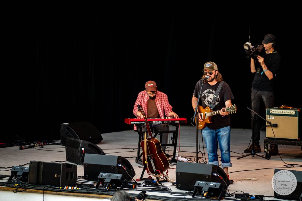 Three musicians perform on stage, one playing keyboard, one on guitar, and one filming with a camera.