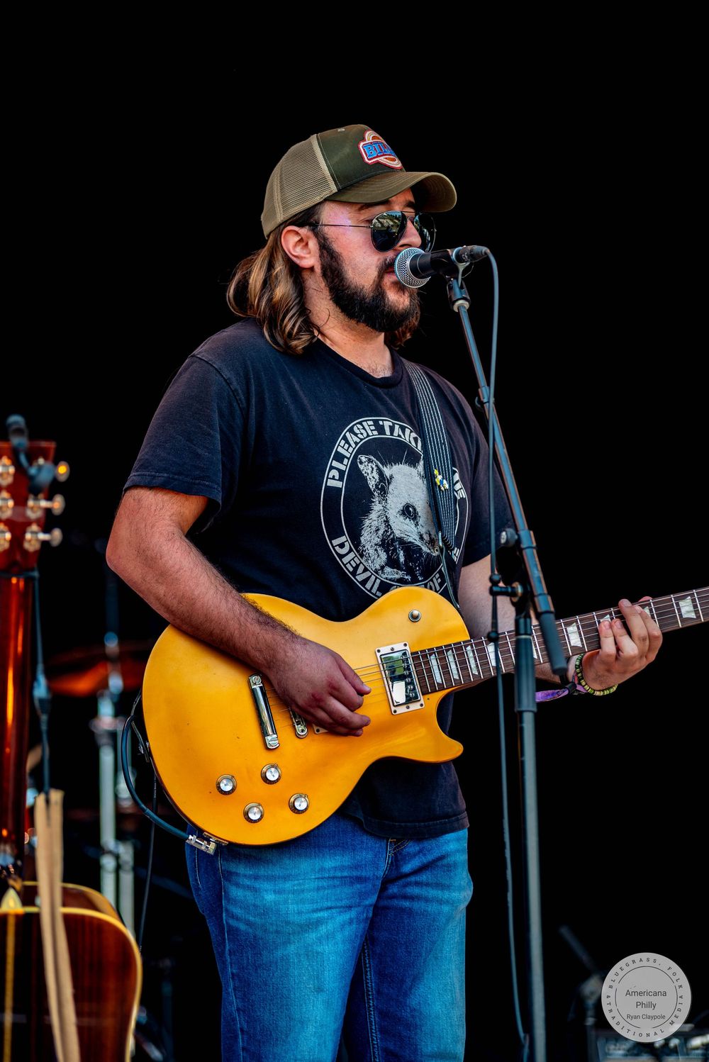 Bearded male guitarist singing into microphone on stage, wearing cap and sunglasses, playing gold Les Paul guitar at outdoor concert.