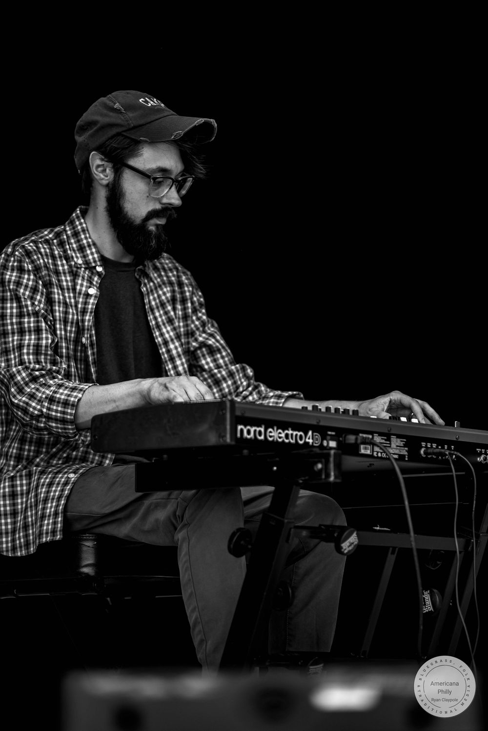 A bearded musician plays a Nord Electro 4 keyboard on stage in a black and white photo.