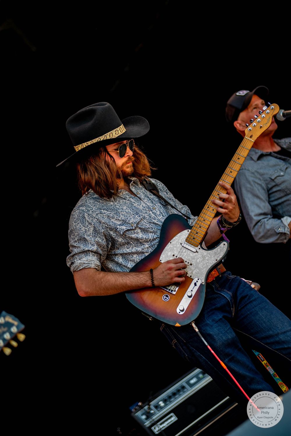 Man in black cowboy hat playing electric guitar on stage with another musician visible in background.