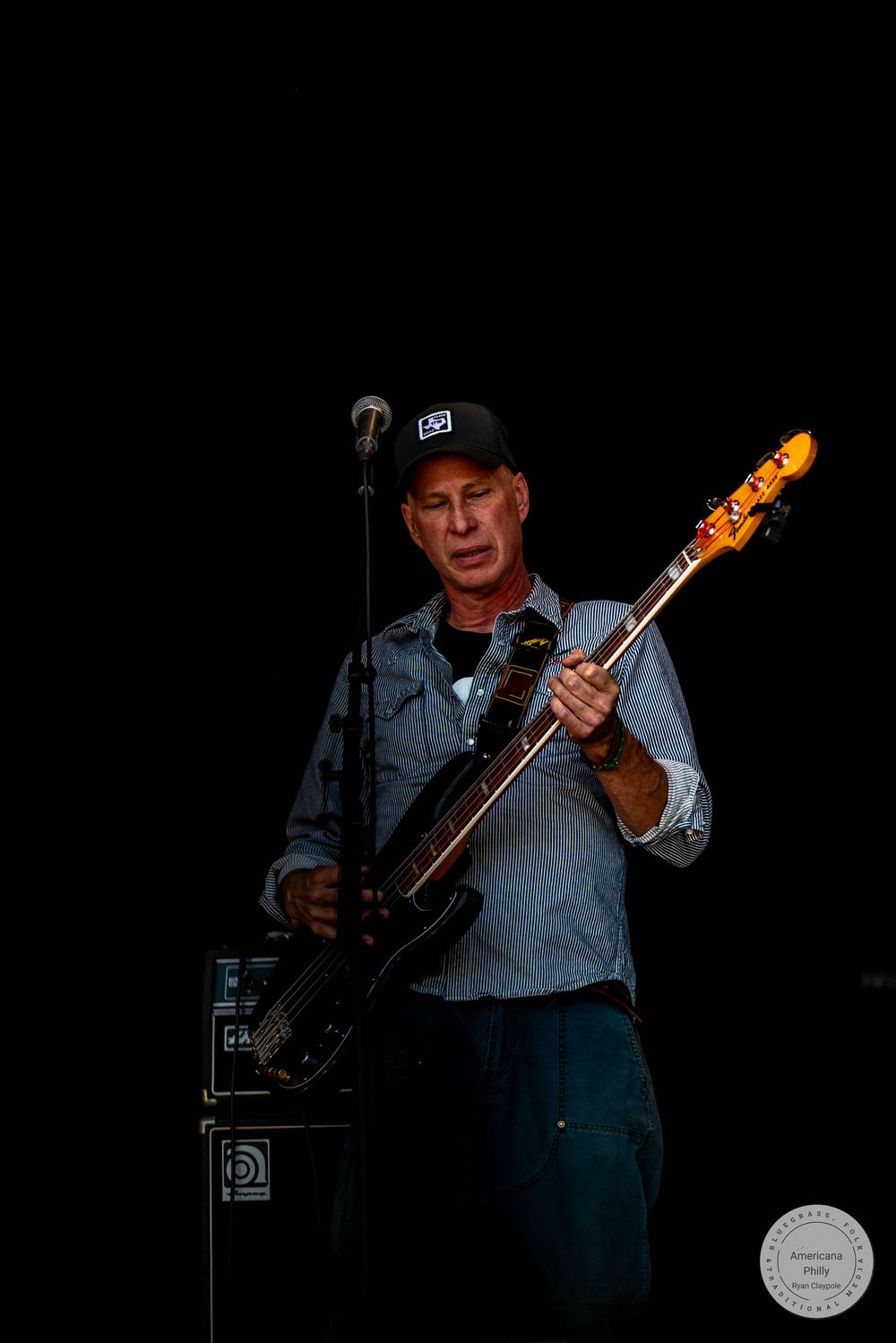 Man playing black bass guitar on stage, wearing a cap and blue shirt, performing live in dark concert setting.