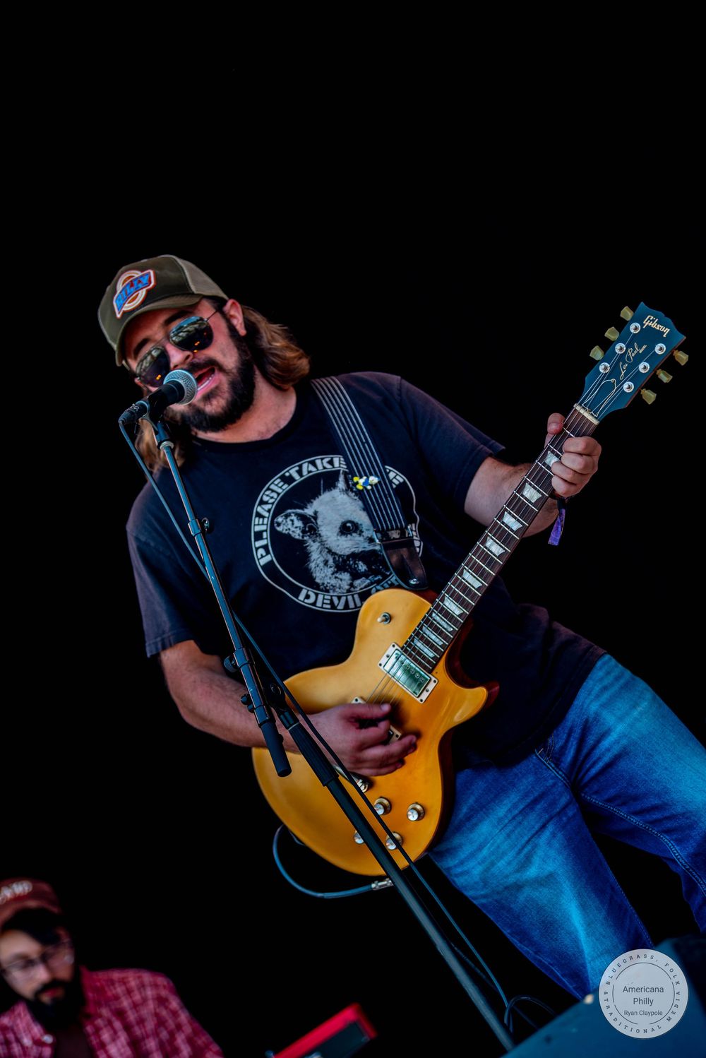 Male guitarist performing on stage, wearing a cap and sunglasses, playing a gold Les Paul guitar while singing into a microphone.