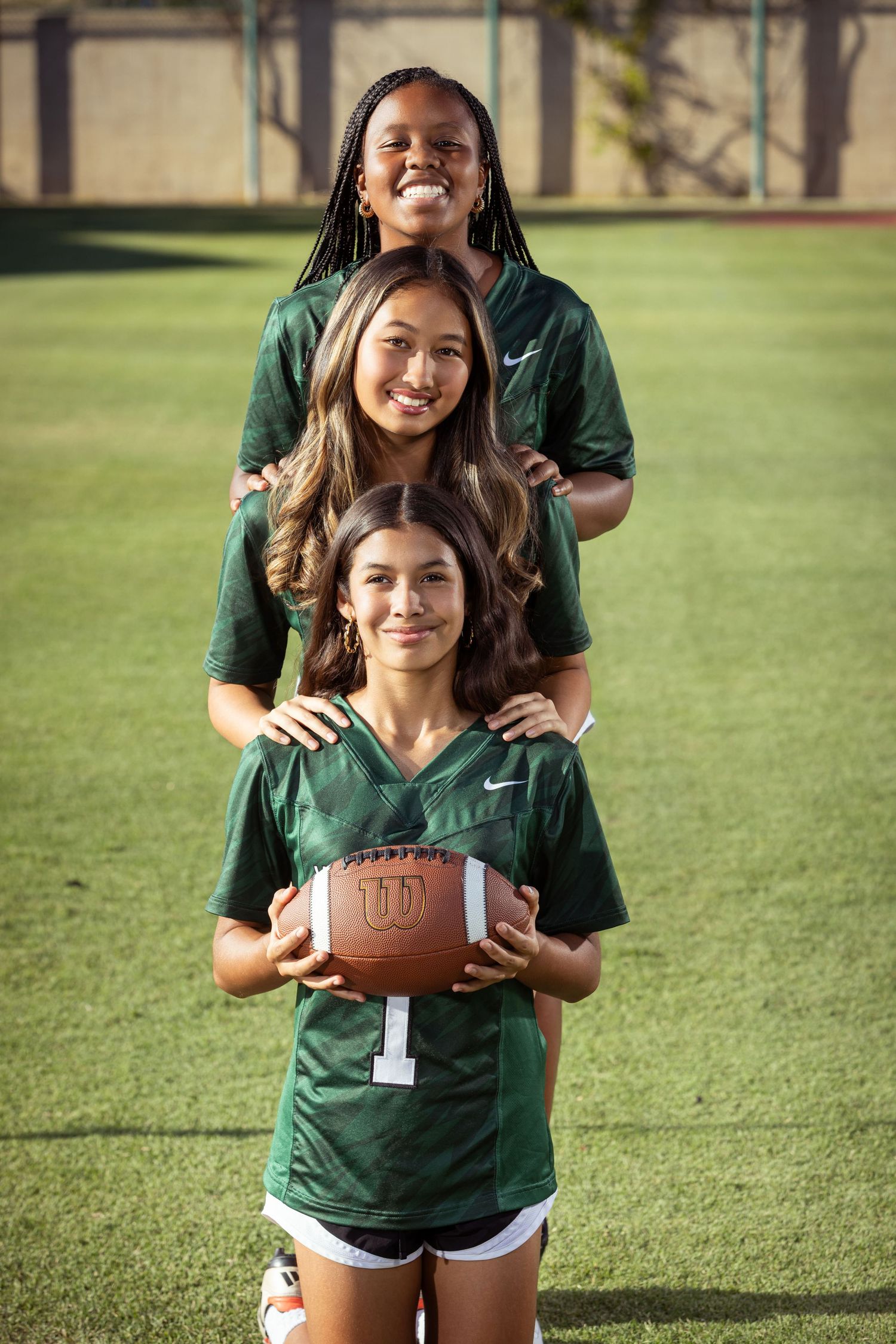 Three girls in green Nike football jerseys pose on a field, with the front girl holding a Wilson football.
