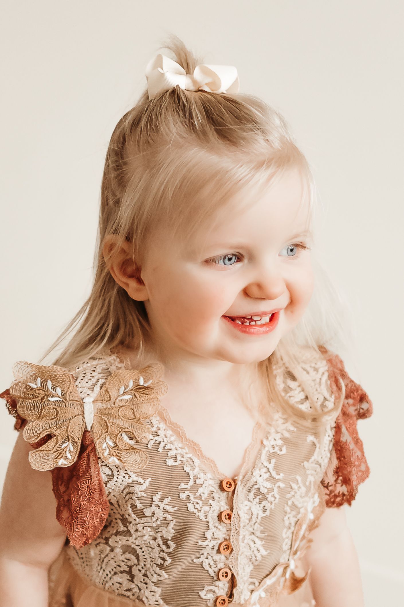 Smiling toddler girl with blonde hair and blue eyes wearing a lace dress with a white bow hair clip.