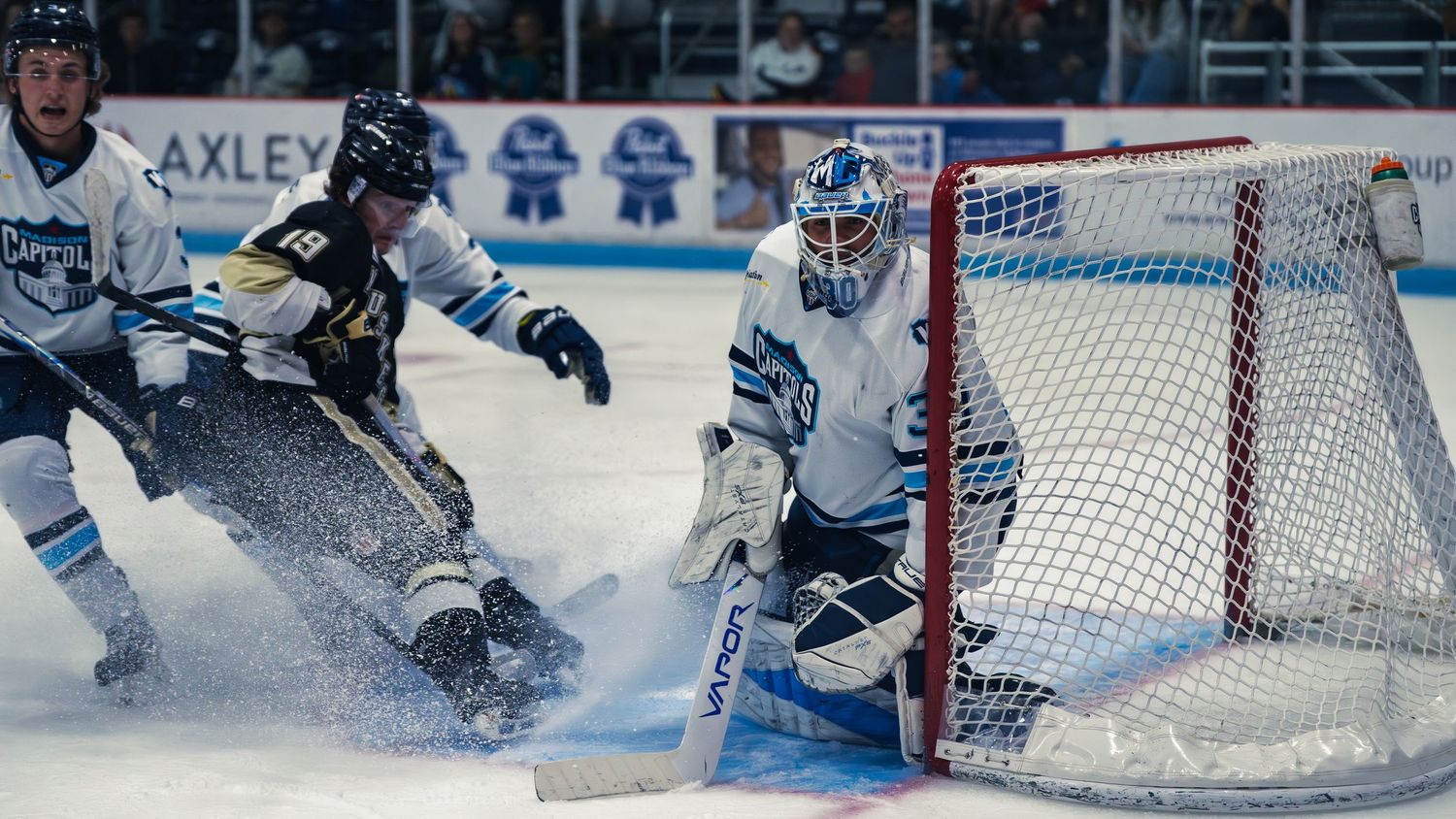 A white Madison Capitols goaltender number 30 hugs the post as a black-and-gold Lumberjacks player number 19 sprays ice at the crease.