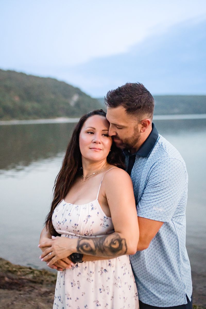 A couple embraces tenderly by a serene lake, the man kissing her head, both sharing a romantic outdoor moment.