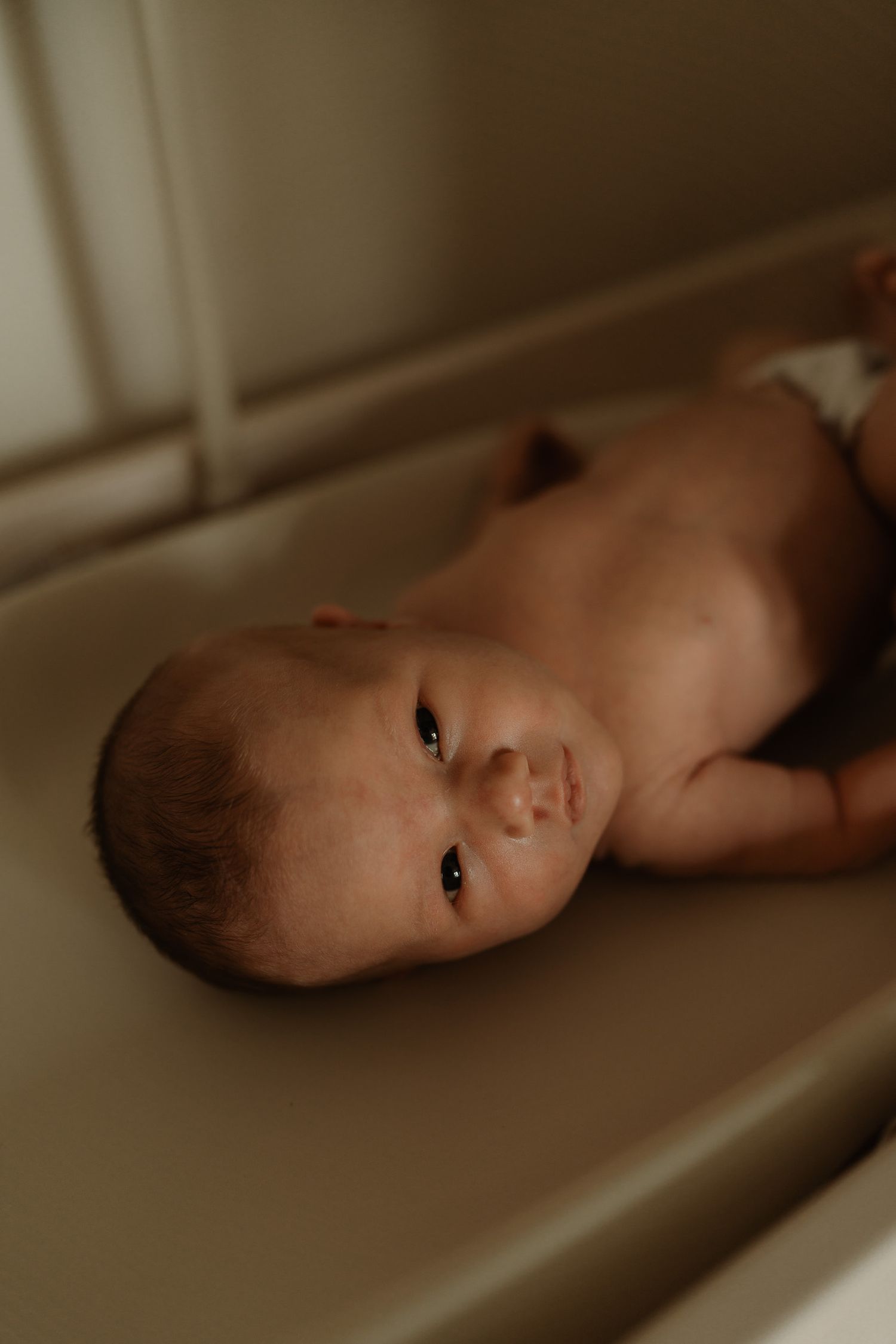 A newborn baby lying on a changing table, looking up with dark eyes, wearing only a diaper.