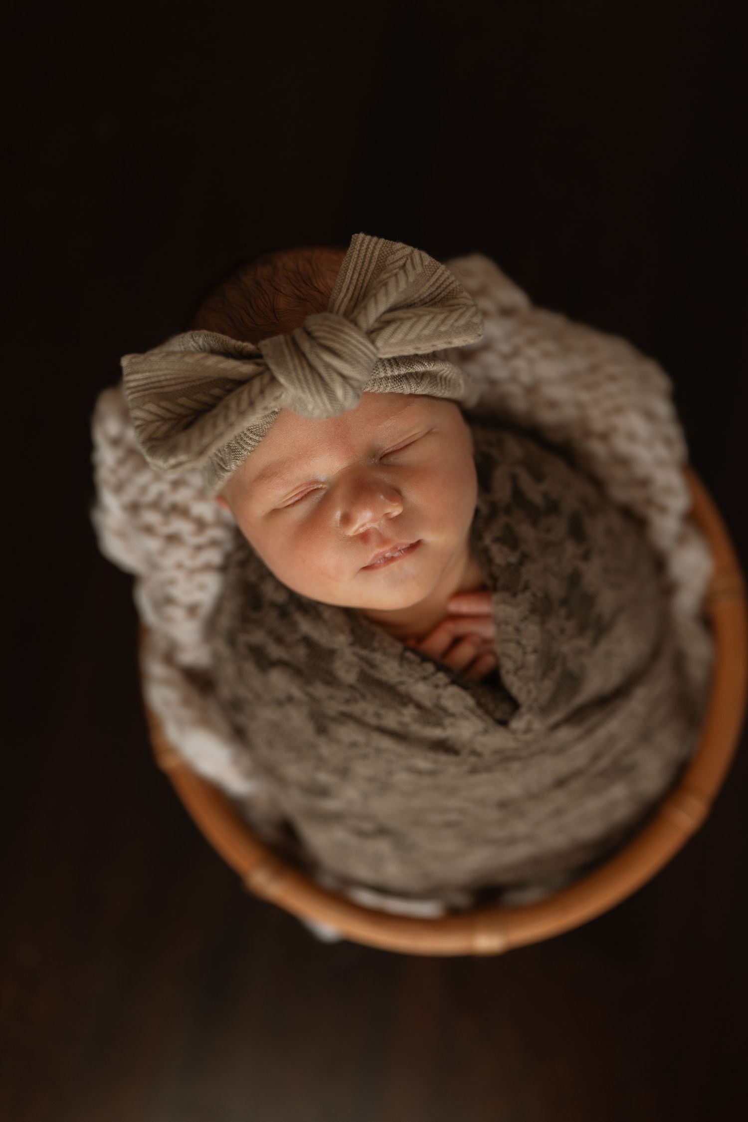 Sleeping newborn baby wrapped in a cozy gray knit blanket with a bow headband, nestled in a wooden basket.