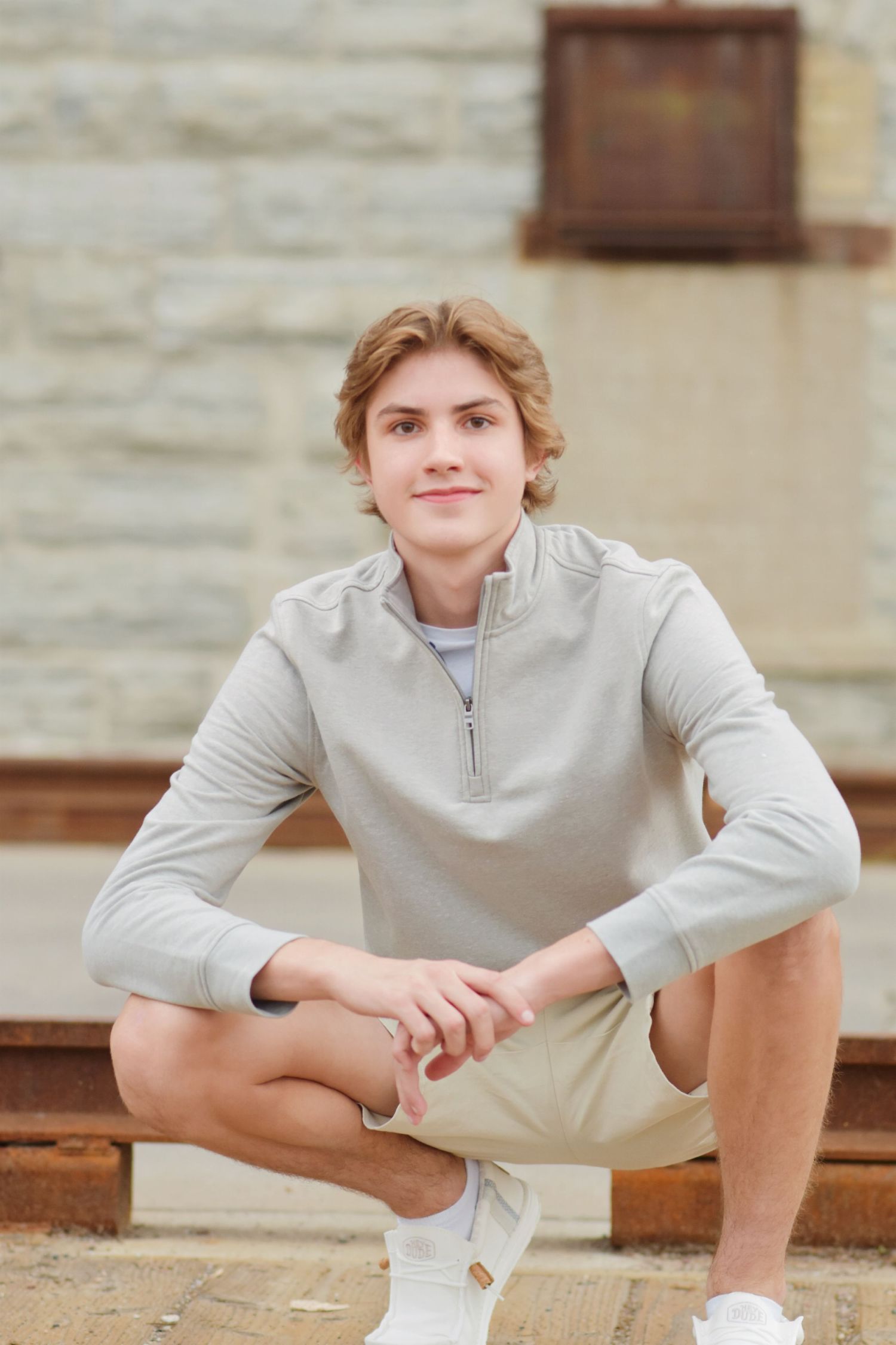 Young man in gray quarter-zip and khaki shorts sitting confidently outdoors near a stone wall.