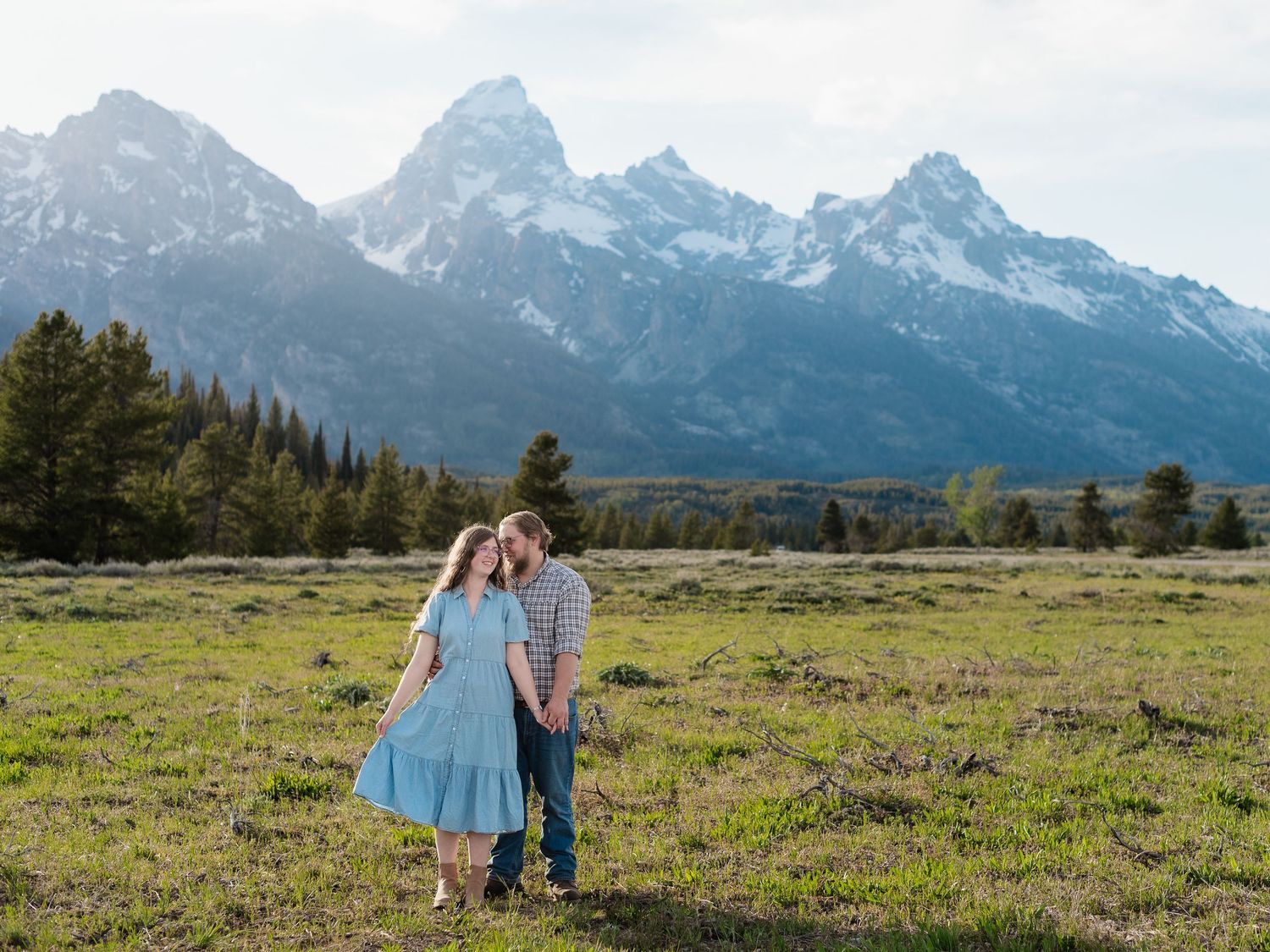 Couple embracing in a meadow with the Grand Teton mountain range in the background on a sunny day.