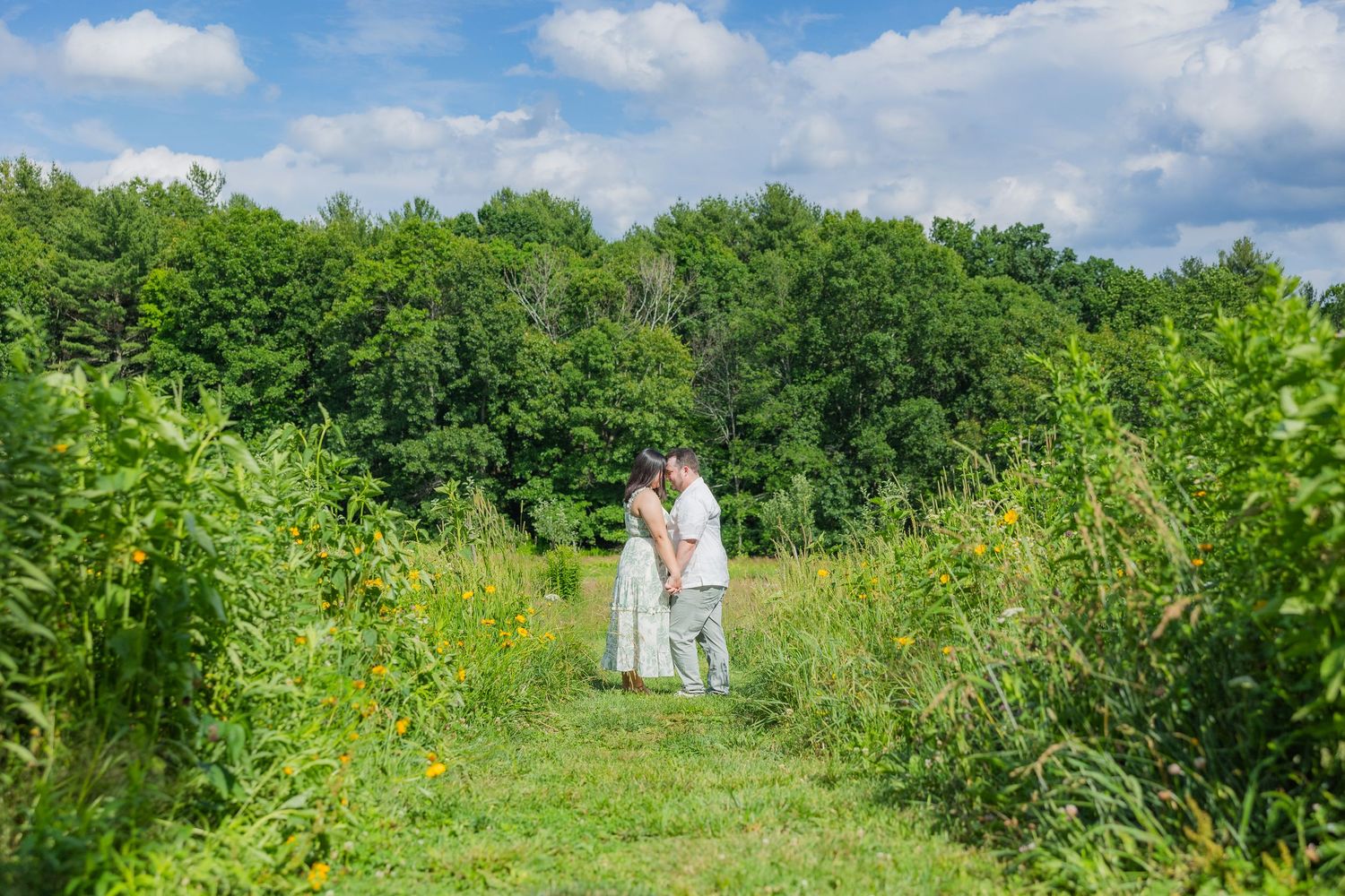 Couple sharing a romantic moment in a lush green meadow surrounded by wildflowers and trees under a blue sky.