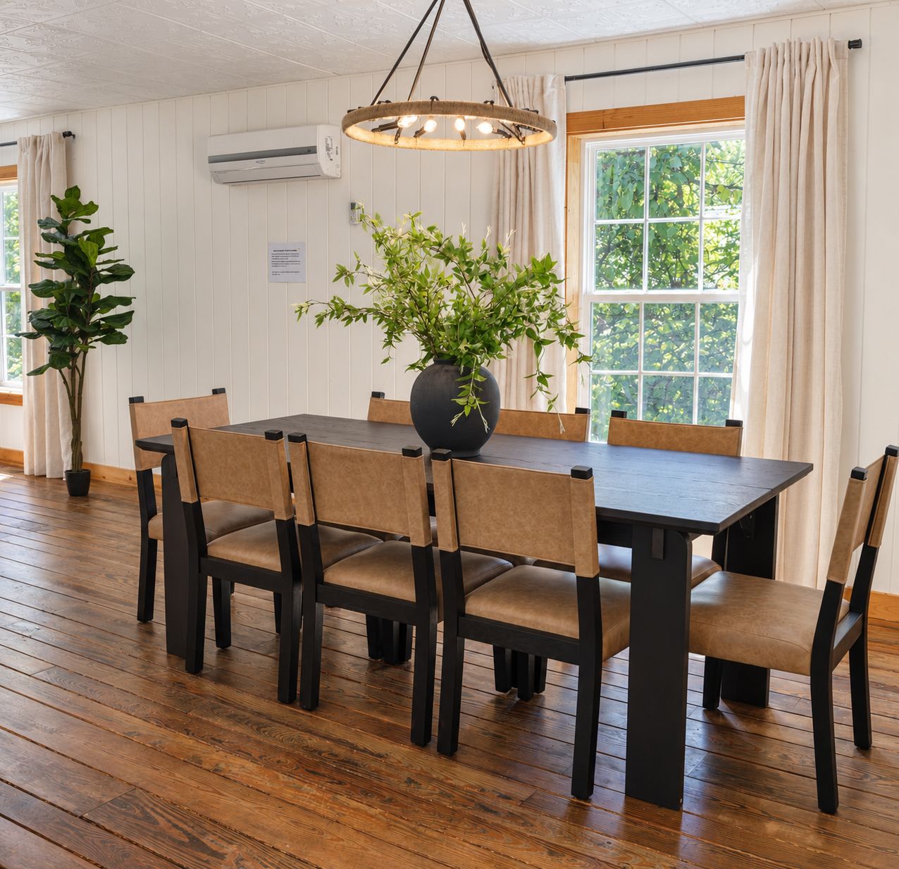 Rustic dining room with dark wood table, woven chairs, hardwood floors, chandelier, and large window with white curtains.