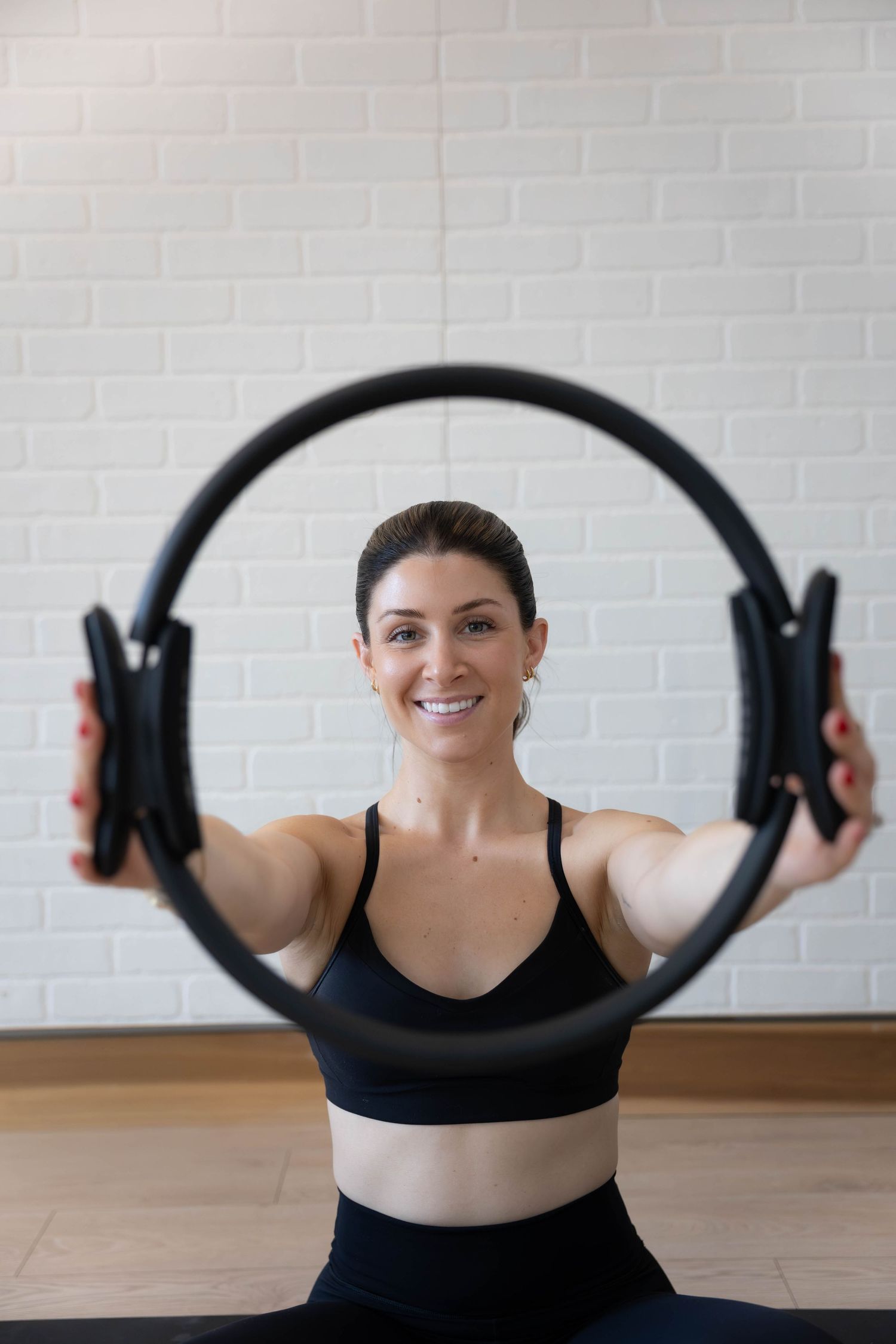 Smiling woman in black activewear holds a Pilates ring in a bright studio with white brick walls.