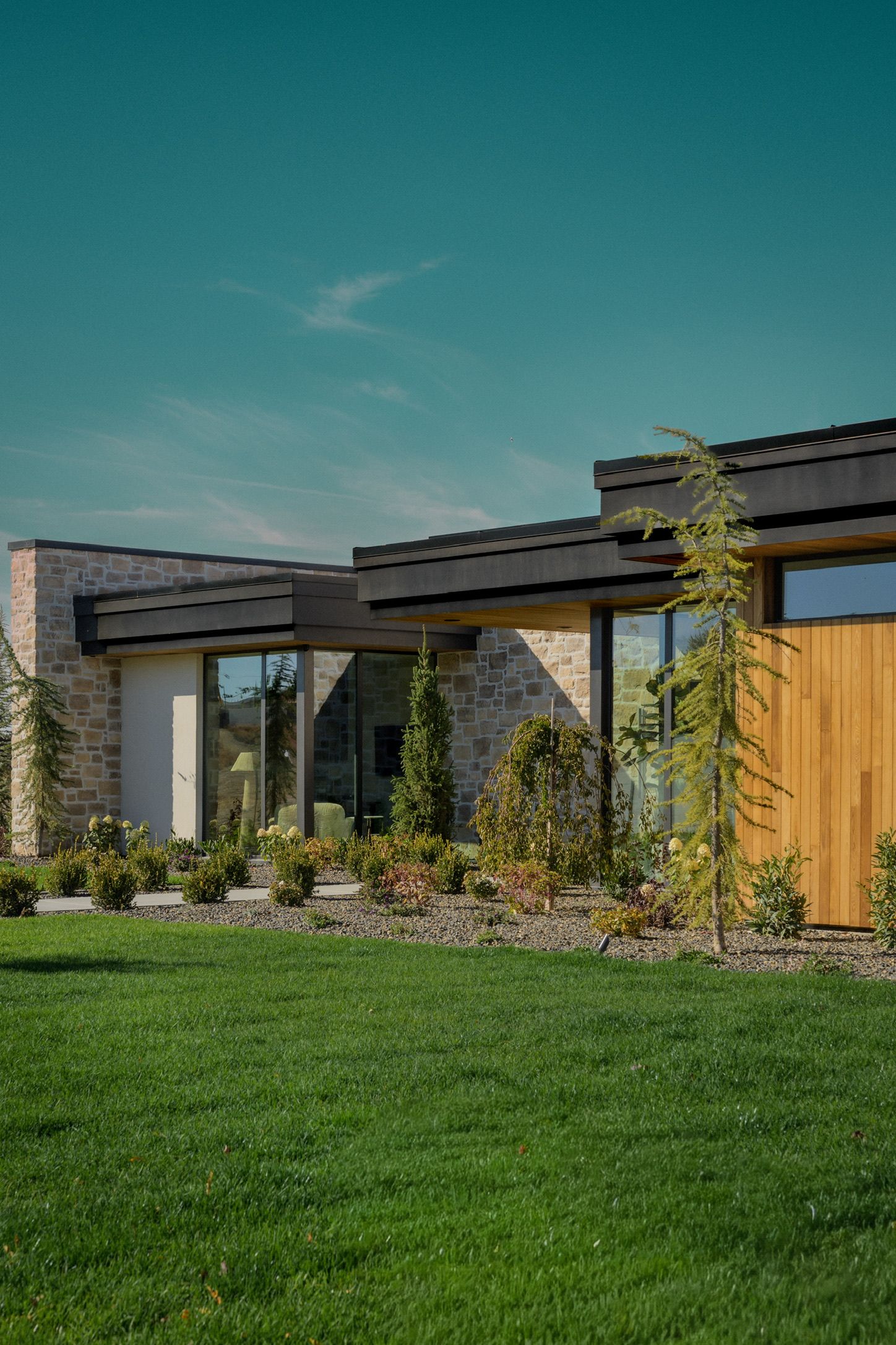 Modern single-story house with stone walls, cedar wood paneling and dark flat roofs, surrounded by a lush green lawn and landscaped garden.