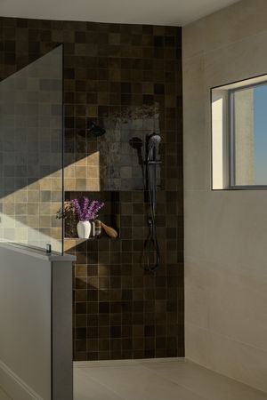 Modern walk-in shower with dark brown mosaic tiles, matte black fixtures, built-in niche shelf, and a glass panel enclosure.
