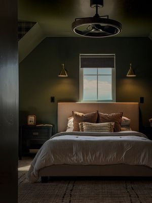 Moody green bedroom with upholstered bed, warm throw pillows, brass wall sconces, and industrial ceiling fan.