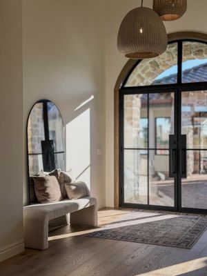 Modern entryway with arched steel door, woven pendant lights, curved bench, tall mirror, and hardwood floors in neutral tones.