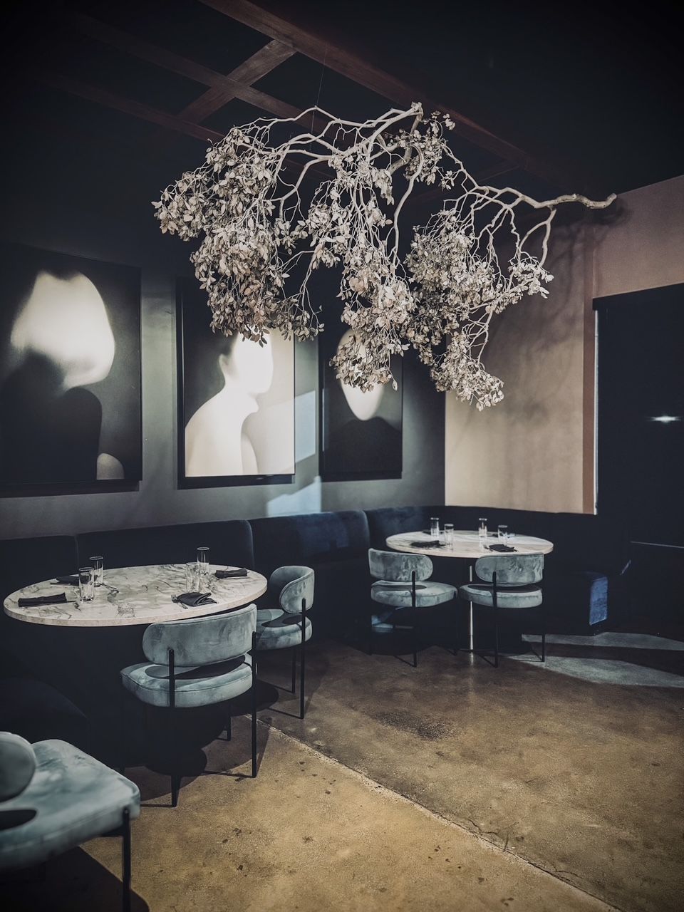 Elegant dark restaurant interior with silver branch installation, teal velvet chairs, marble tables, and moody artwork on walls.