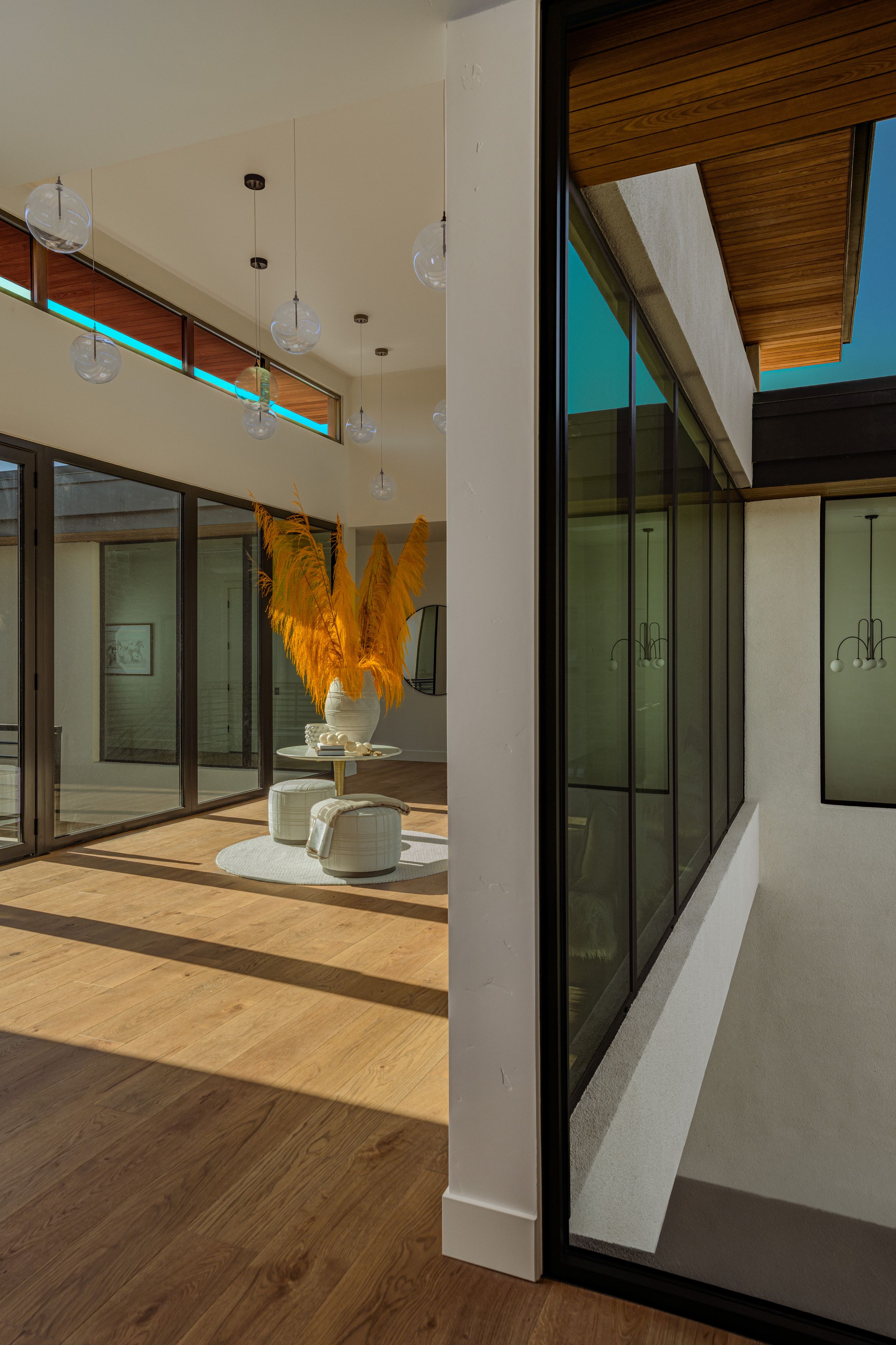 Modern interior hallway with floor-to-ceiling glass walls, light hardwood flooring, pendant lights and golden pampas grass on a round table.