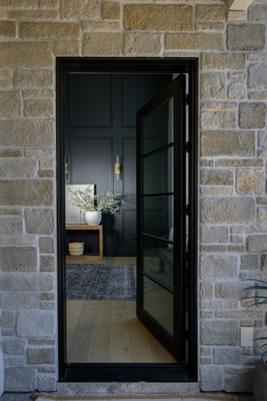 Black steel-framed glass pivot front door set in natural stone wall opening to dark elegant entryway.