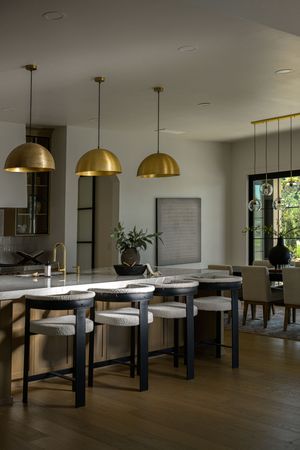 Modern kitchen with gold dome pendant lights, black bar stools, marble island, and open dining area.