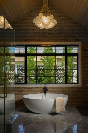 Luxurious bathroom with freestanding soaking tub, brick walls, bubble chandelier, and black-framed window.