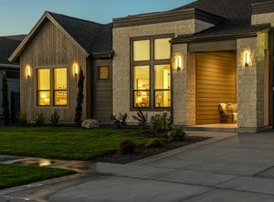 Modern farmhouse exterior at dusk with warm glowing windows and elegant wall sconces illuminating the stone and wood facade.